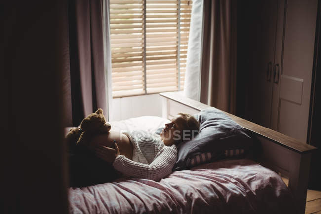 Беременная женщина держит плюшевого мишку на животе, пока спит в спальне дома — стоковое фото