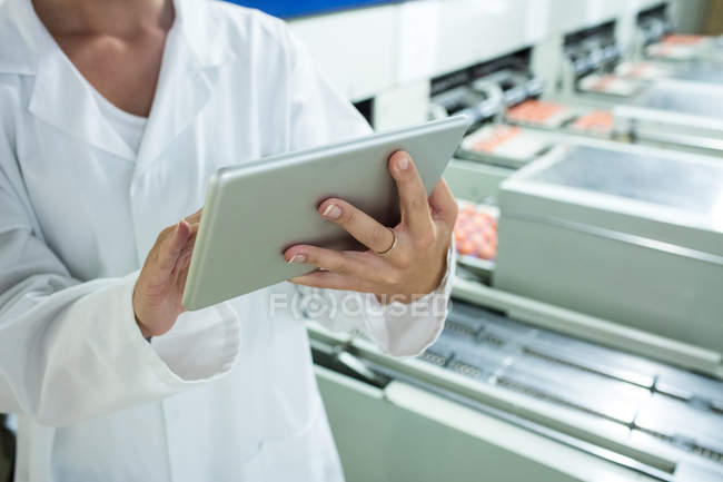 Sección media del personal femenino que utiliza tableta digital junto a la línea de producción en la fábrica de huevos - foto de stock