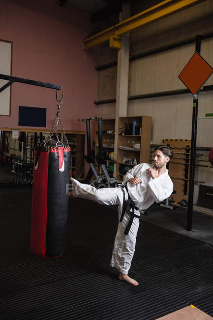 Vue grand angle de l'homme pratiquant le karaté avec sac de boxe dans le studio de fitness — Photo de stock