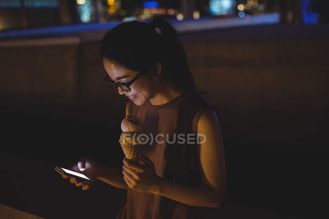 Jeune femme utilisant un téléphone portable tout en ayant de la crème glacée la nuit — Photo de stock