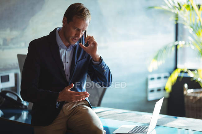 Hombre de negocios hablando en el teléfono móvil mientras usa el ordenador portátil en la oficina - foto de stock