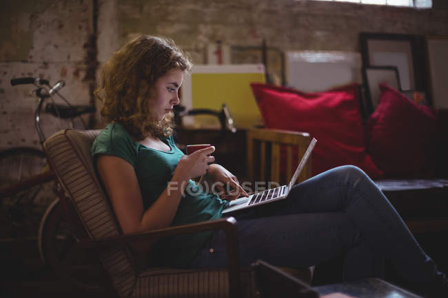 Mecânico bonito usando laptop enquanto toma café na oficina — Fotografia de Stock