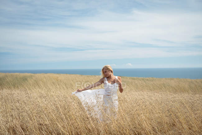 Беззаботная блондинка в белом платье стоит в поле — стоковое фото