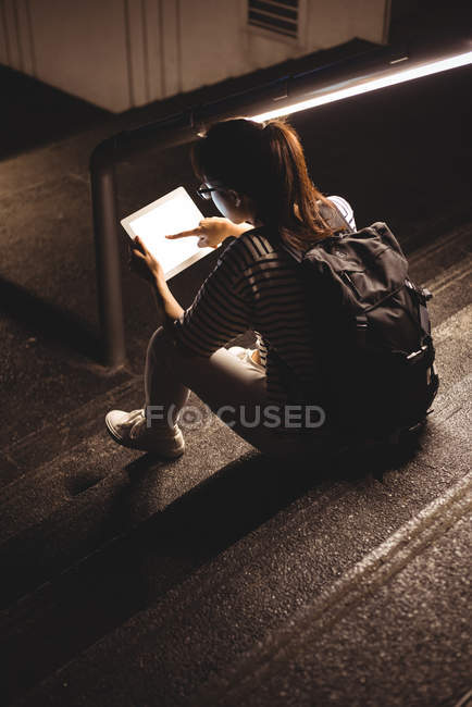 Вид сзади на молодую женщину, сидящую на лестнице и использующую цифровые планшеты ночью — стоковое фото
