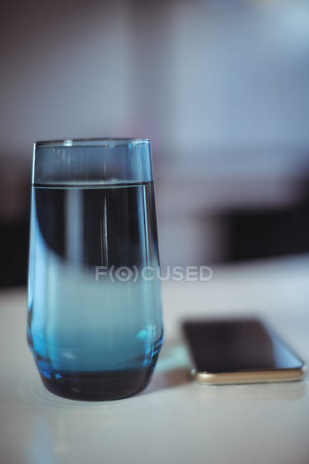 Стакан воды и мобильный телефон на столе в офисе — стоковое фото