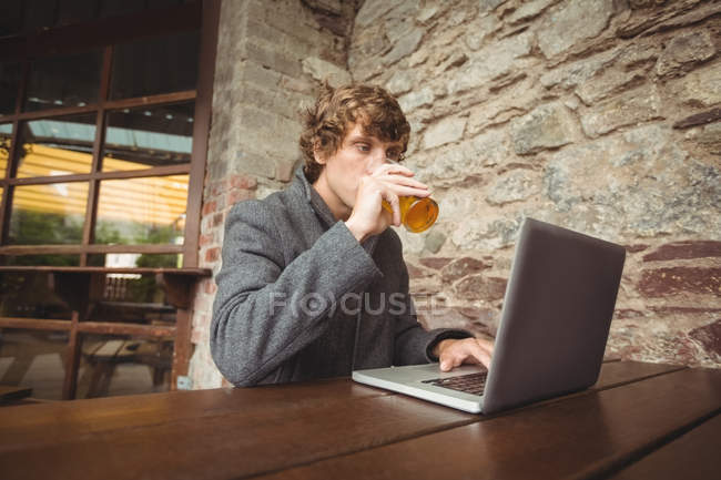 Partie médiane de l'homme tenant un verre de bière et utilisant un ordinateur portable au bar — Photo de stock