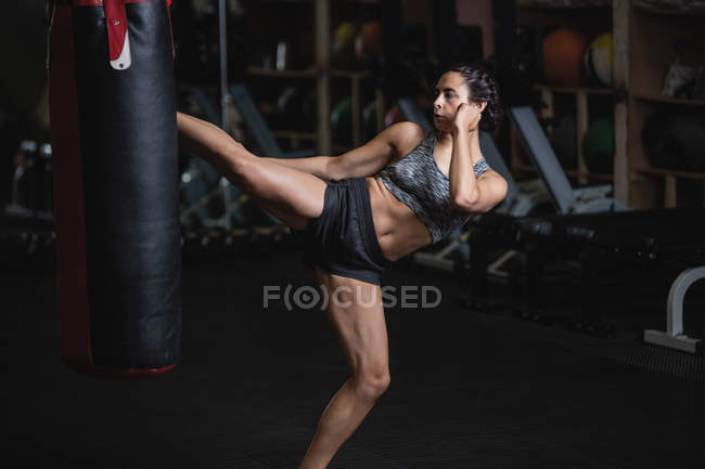 Vista lateral do boxer feminino chutando saco de perfuração no estúdio de fitness — Fotografia de Stock