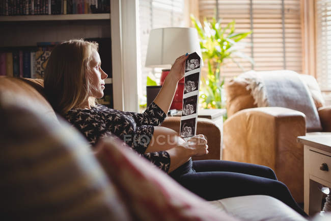 Vista lateral da mulher grávida olhando para uma ultrassonografia na sala de estar em casa — Fotografia de Stock