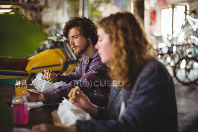 Pareja sentada en la mesa y comiendo sándwich en la tienda de bicicletas - foto de stock