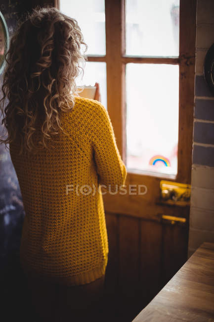 Жінка дивиться крізь вікно, маючи каву на кухні вдома — стокове фото