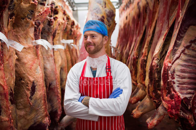 Carnicero sonriente de pie con los brazos cruzados en la sala de almacenamiento de carne en la carnicería - foto de stock
