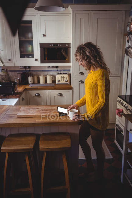 Schöne Frau mit digitalem Tablet, während sie zu Hause in der Küche Kaffee trinkt — Stockfoto