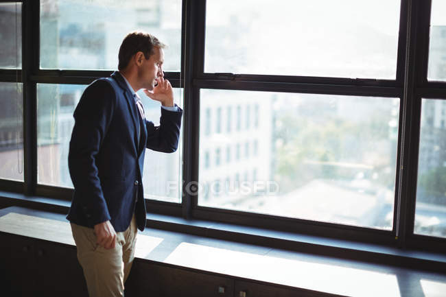 Businessman looking through window while talking on the phone in office — Stock Photo