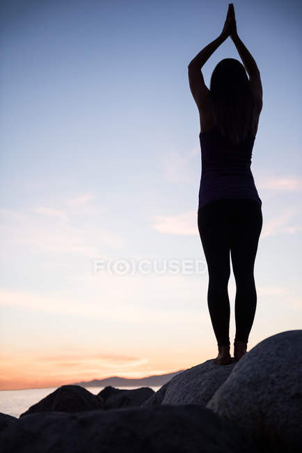 Rückansicht einer Frau, die in der Abenddämmerung Yoga auf einem Felsen praktiziert — Stockfoto