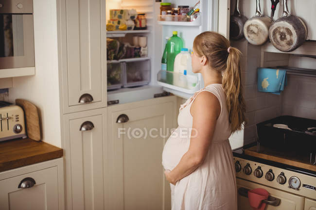 Беременная женщина ищет еду в холодильнике на кухне — стоковое фото