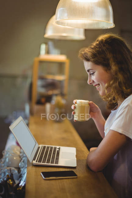 Beautiful mechanic having coffee while using laptop at counter in workshop — Stock Photo