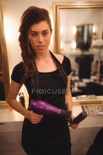 Female hairdresser holding hair dryer machine and hairbrush at saloon — Stock Photo