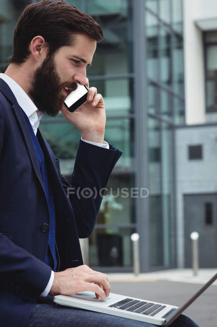 Homme d'affaires parlant sur le téléphone mobile tout en utilisant ordinateur portable côté extérieur — Photo de stock
