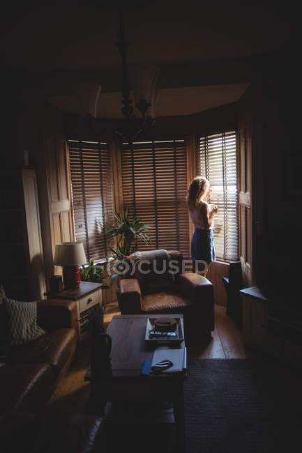 Femme regardant par la fenêtre dans le salon à la maison — Photo de stock