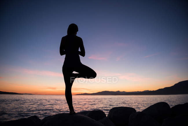 Vista posteriore della donna che pratica yoga sulla roccia al tramonto — Foto stock