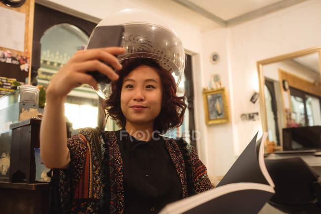Stylish woman taking a selfie while sitting under a hairdryer at hair salon — Stock Photo