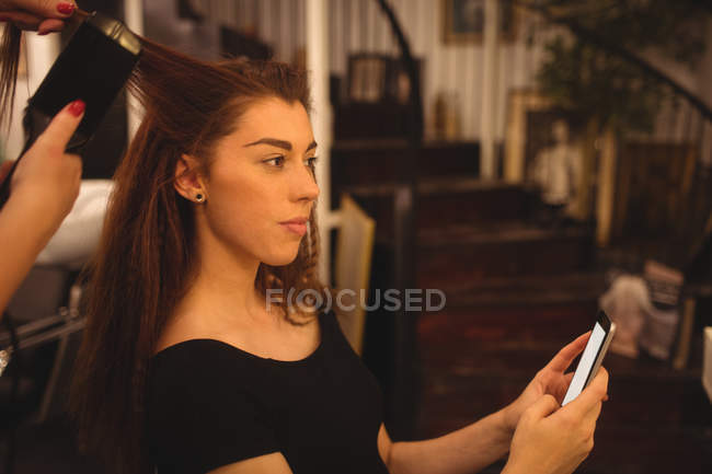 Woman using mobile phone while getting hair straightened at hair saloon — Stock Photo