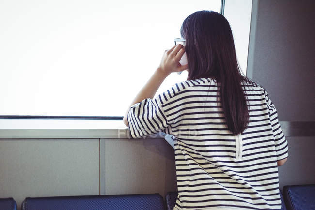 Вид ззаду жінки, що дивиться через вікно під час розмови на мобільному телефоні — стокове фото