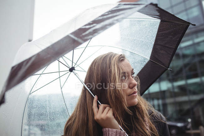 Hermosa mujer sosteniendo paraguas durante la temporada de lluvias en la calle y mirando hacia arriba - foto de stock