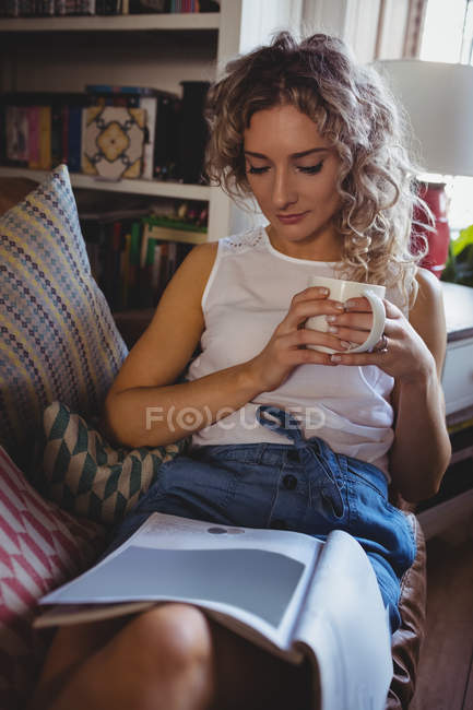 Schöne Frau liest Magazin, während sie zu Hause im Wohnzimmer Kaffee trinkt — Stockfoto