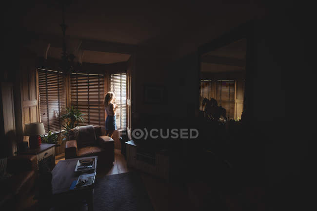Mulher olhando através da janela na sala de estar em casa — Fotografia de Stock