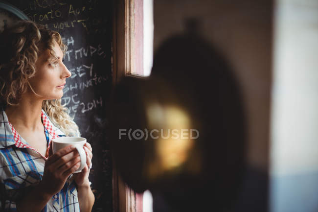 Красива жінка дивиться крізь вікно, маючи каву вдома — стокове фото