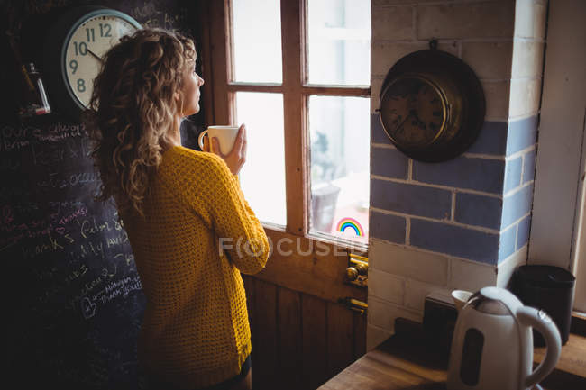 Frau schaut durch Fenster, während sie zu Hause in der Küche Kaffee trinkt — Stockfoto
