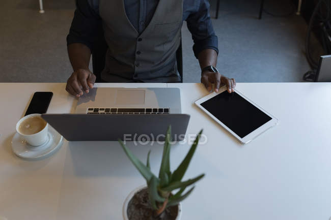 Sección media del hombre de negocios usando el ordenador portátil y la tableta digital en la oficina - foto de stock