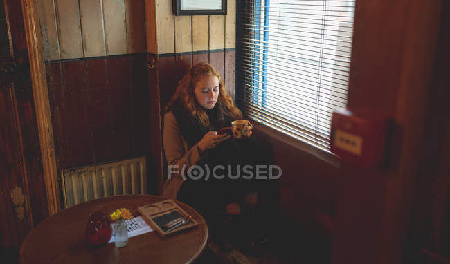 Mulher ruiva usando telefone celular enquanto toma café no café — Fotografia de Stock