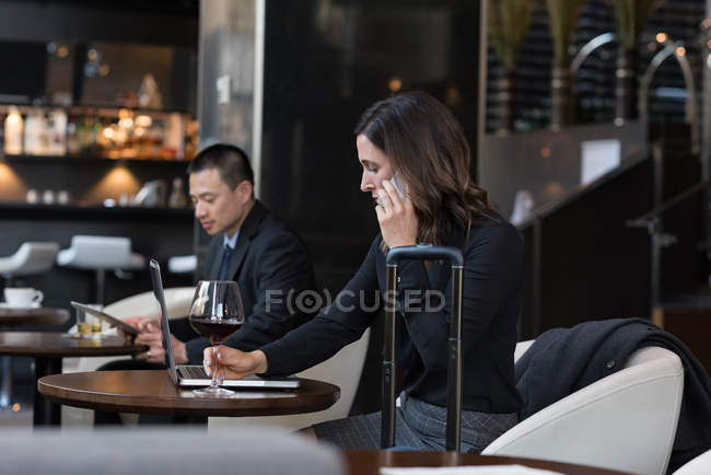 Mujer de negocios tomando vino tinto mientras habla por teléfono móvil en el hotel - foto de stock