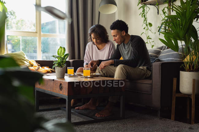 Пара використовує мобільний телефон, маючи їжу на дивані у вітальні вдома — стокове фото