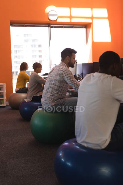 Ejecutivos de negocios trabajando en el escritorio mientras están sentados en la pelota de ejercicio en la oficina - foto de stock