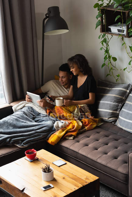 Пара с помощью мобильного телефона и цифрового планшета во время кофе на диване в гостиной на дому — стоковое фото