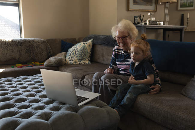Abuela y nieta haciendo videollamada en el ordenador portátil en la sala de estar en casa - foto de stock