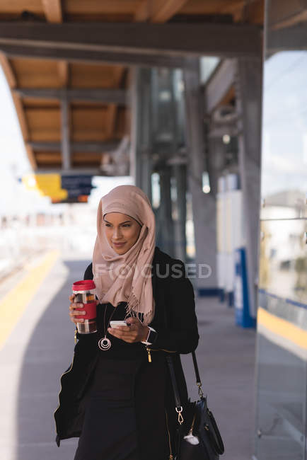 Hijab mujer usando el teléfono móvil mientras toma café en la plataforma en la estación de tren - foto de stock