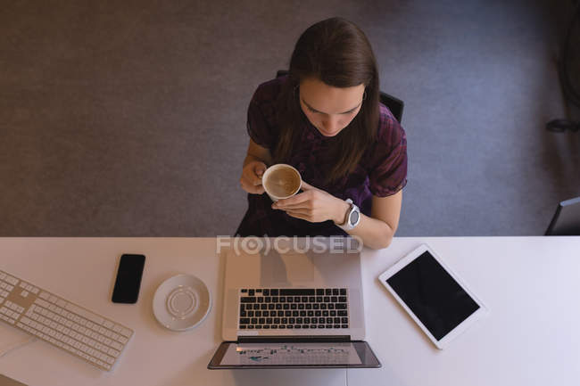 Belle femme d'affaires prenant un café tout en utilisant un ordinateur portable au bureau — Photo de stock