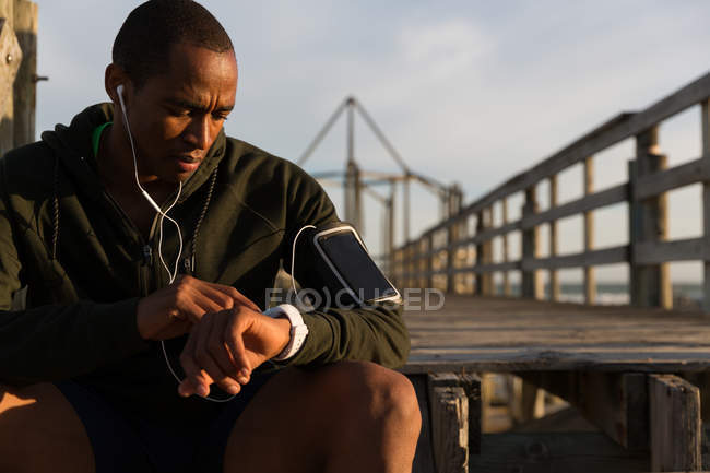 Athlète masculin utilisant sa smartwatch sur la jetée à la plage — Photo de stock