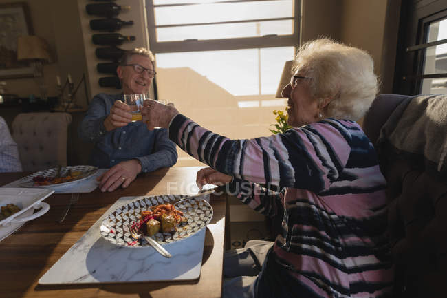 Senior pareja tostando vaso de jugo en la mesa de comedor en casa - foto de stock
