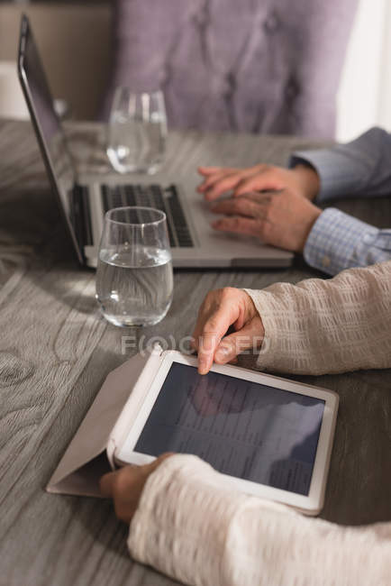 Primer plano de la pareja de personas mayores utilizando el ordenador portátil y la tableta digital en la mesa de comedor en casa - foto de stock
