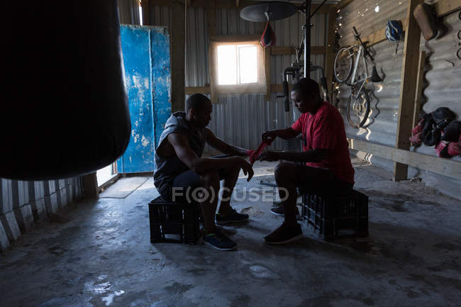 Trainer assisting male boxer in tying hand wrap on hand in fitness studio — Stock Photo