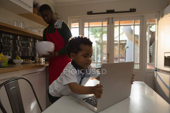 Сын с ноутбуком, пока отец готовит еду на кухне дома — стоковое фото