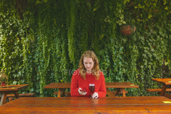 Рыжая женщина, пользующаяся мобильным телефоном за пивом в кафе на открытом воздухе — стоковое фото