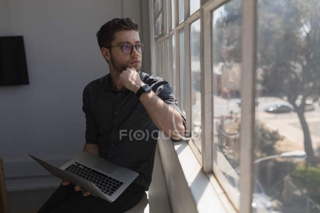 Male executive looking through window while using laptop in office — Stock Photo