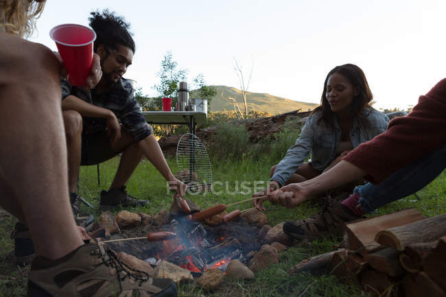 Grupo de amigos assar salsicha na fogueira no acampamento — Fotografia de Stock