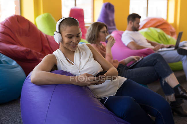 Business executive sorridente che ascolta musica sul cellulare mentre siede sul sacchetto di fagioli — Foto stock